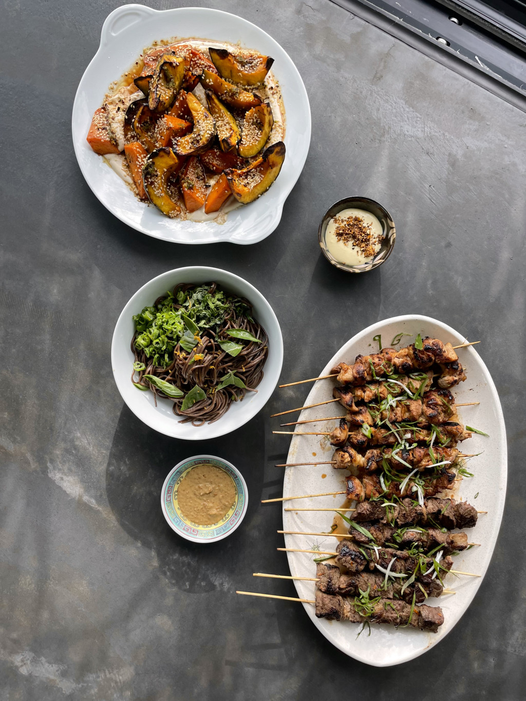 A spread of colorful dishes on a dark grey countertop, including roasted squash and grilled chicken skewers